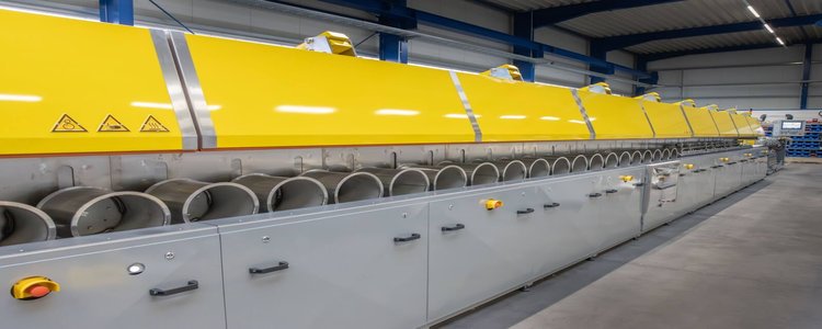 Die zur Besichtigung verfügbare Salzbad-Vulkanisationsanlage: eine lange, glänzende Maschine in einer großen Halle. Die Maschine ist in der oberen Hälfte Gelb und in der unteren Hälfte Grau lackiert.