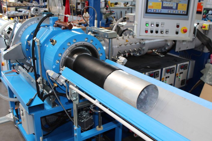An extruder with a large extrusion head is used to coat a metal pipe with rubber.